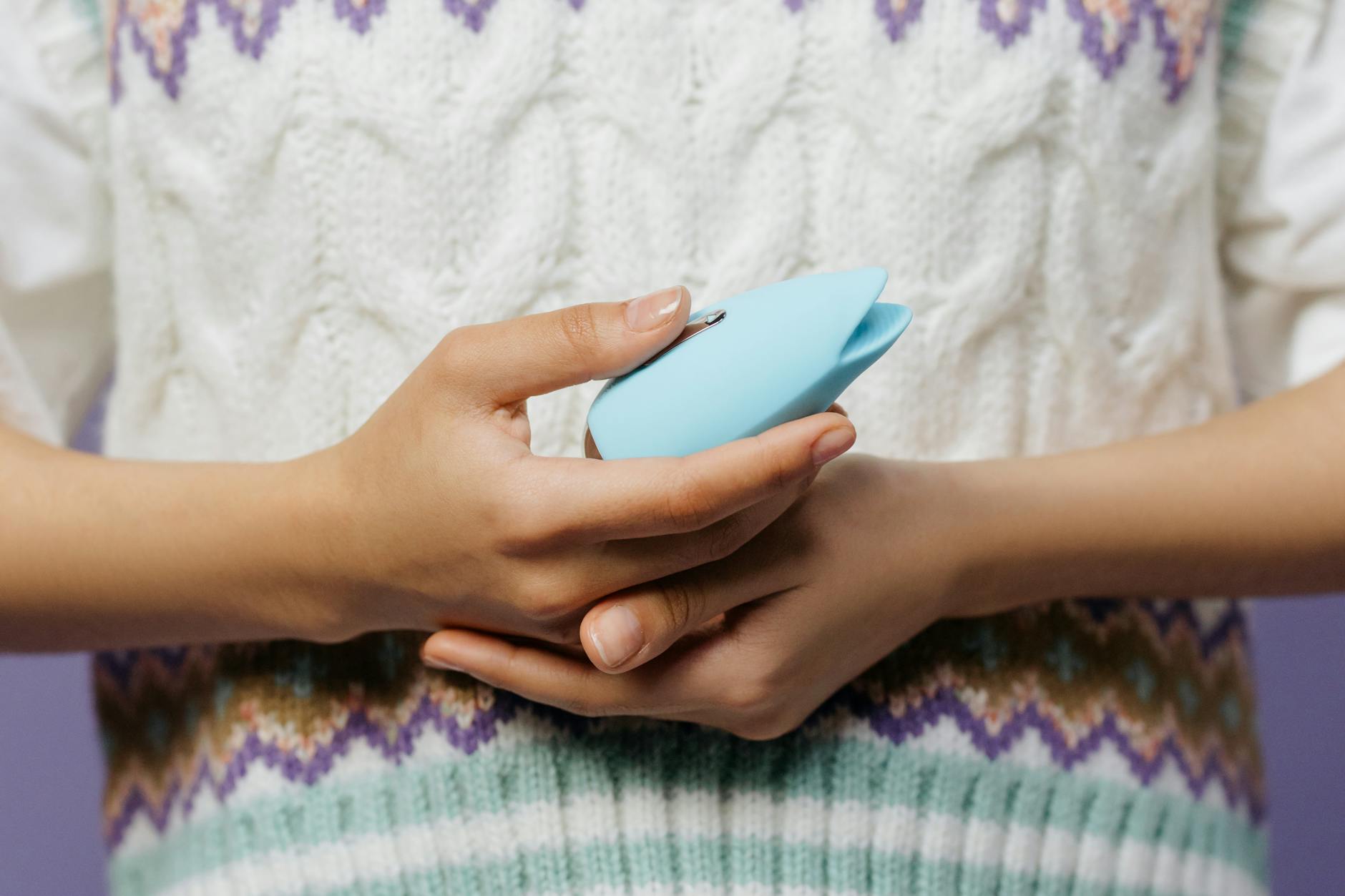 Close-up of hands holding a blue clitoral vibrator against a knitted sweater