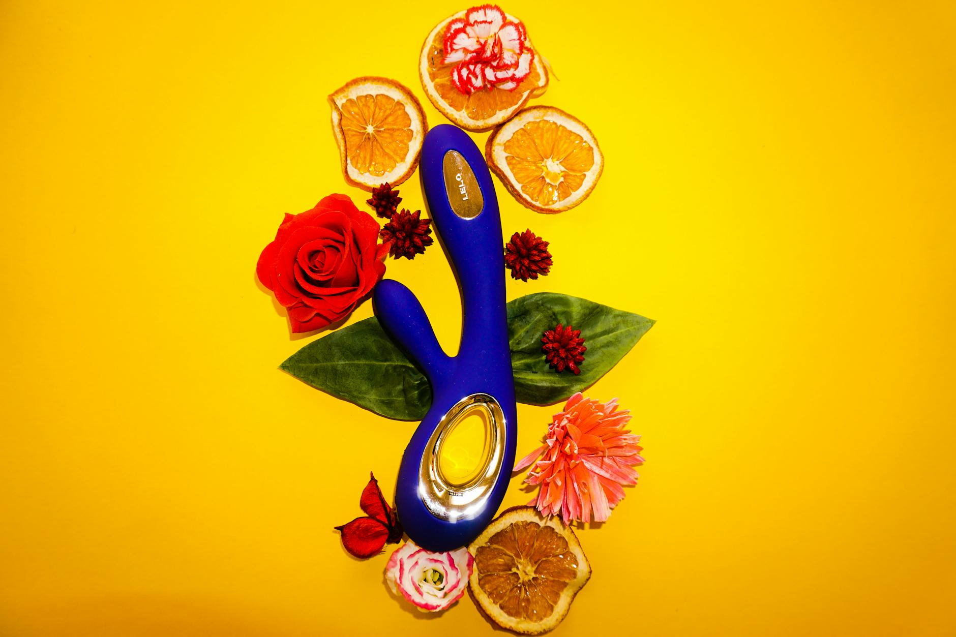 A blue lemon vibrator surrounded by fresh flowers and citrus slices on a bright yellow background.